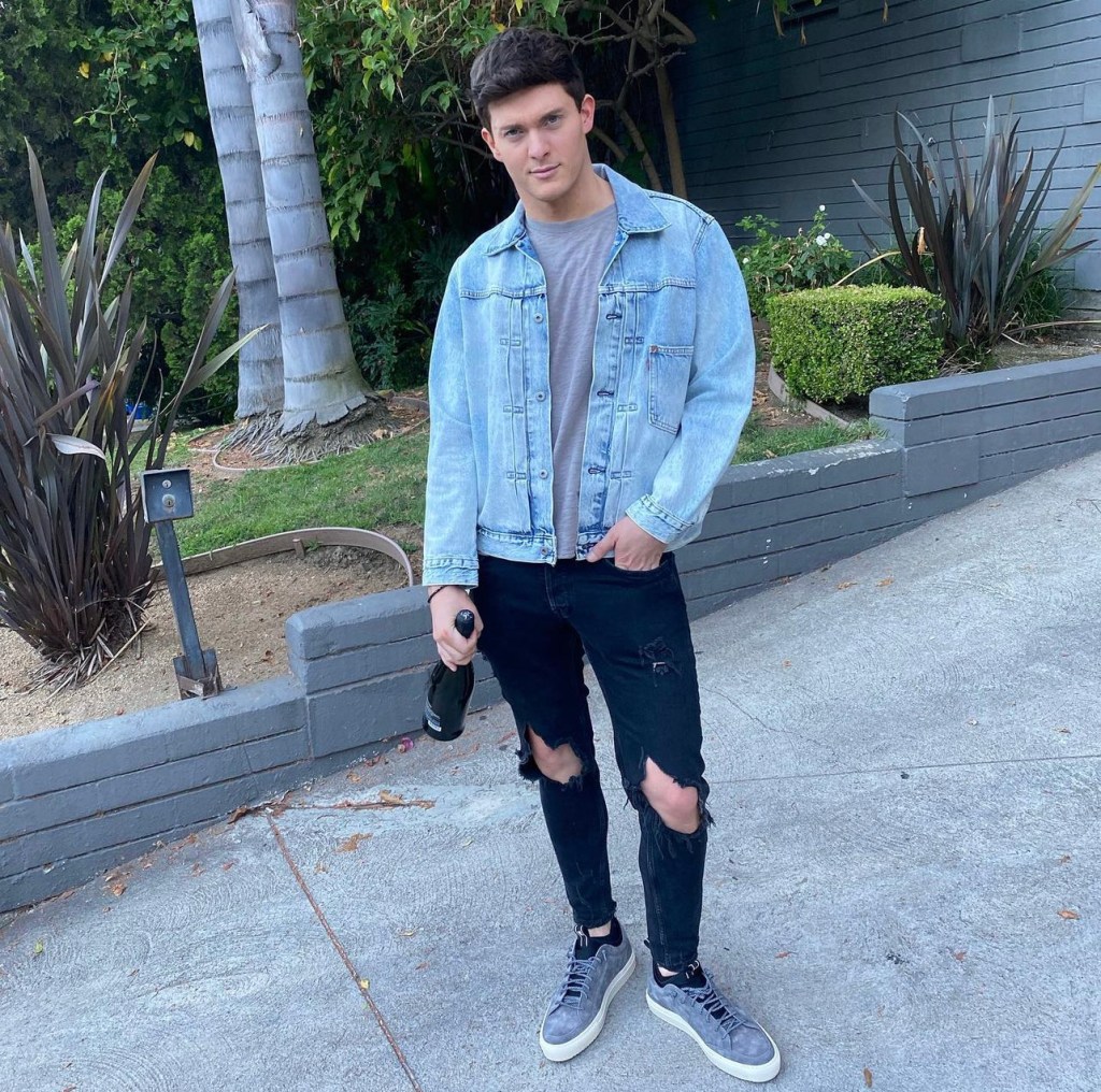 Vanderpump Rules spinoff star Zack Wickham poses for photo in driveway wearing ripped black jeans and jean jacket with gray shirt while holding bottle of champagne